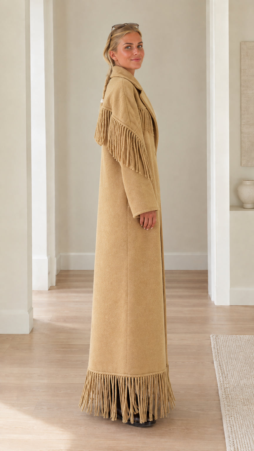 Side profile view of woman wearing long camel overcoat with shoulder and hem fringe