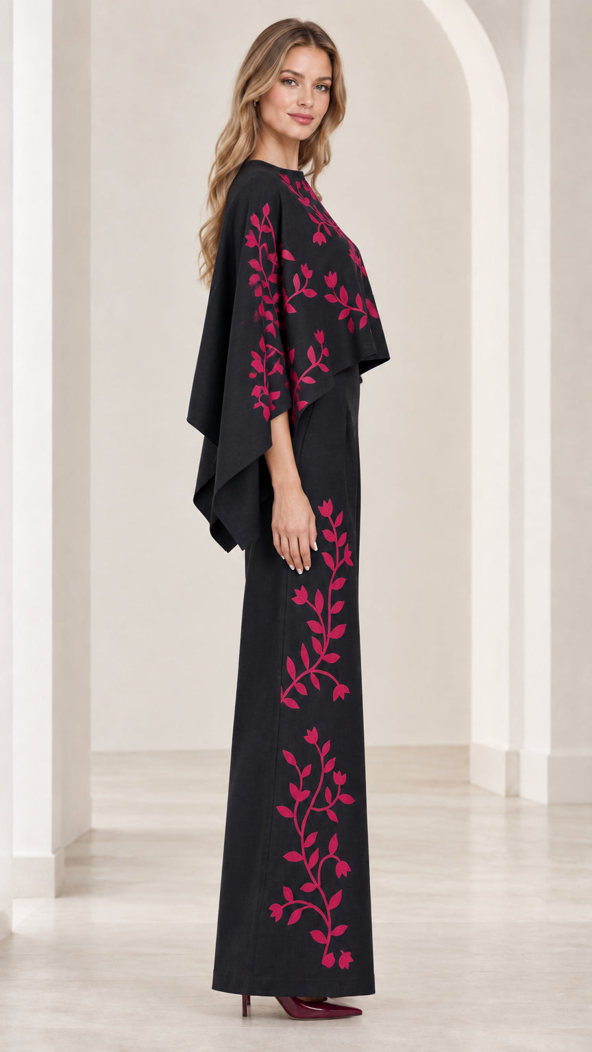 Side profile view of black embroidered cape jumpsuit showing draped capelet